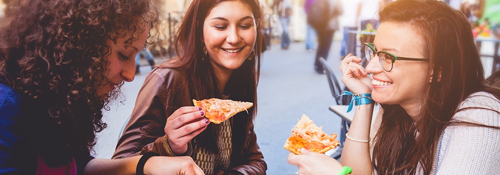 Three young women smiling and eating pizza outdoors